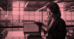 Técnica revisando datos en una tablet en una sala de servidores, con un portátil abierto y servidores activos al fondo, bajo una luz cálida rosada.