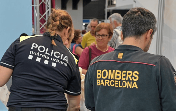 Una agente de la Guardia Urbana y un bombero de Barcelona, de espaldas, atienden a visitantes en un estand